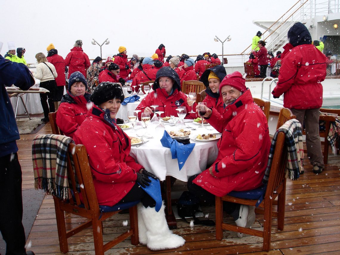 Eine Gruppe von Menschen mit roten Winterjacken sitzt an einer mit weißem Tuch bedeckten Tafel bei einem Essen. Zwei der fünf Personen halten prostend ihre Weingläser Richtung Kamera. Das Essen findet auf eine Schiffsdeck statt. Das Bild ist Teil eines Beitrags über Fleisch und Gemüse.