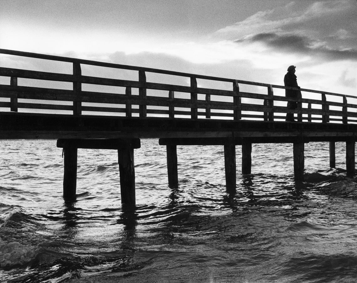 Eine einsame Frau geht über einen Steg und blickt auf einen See.