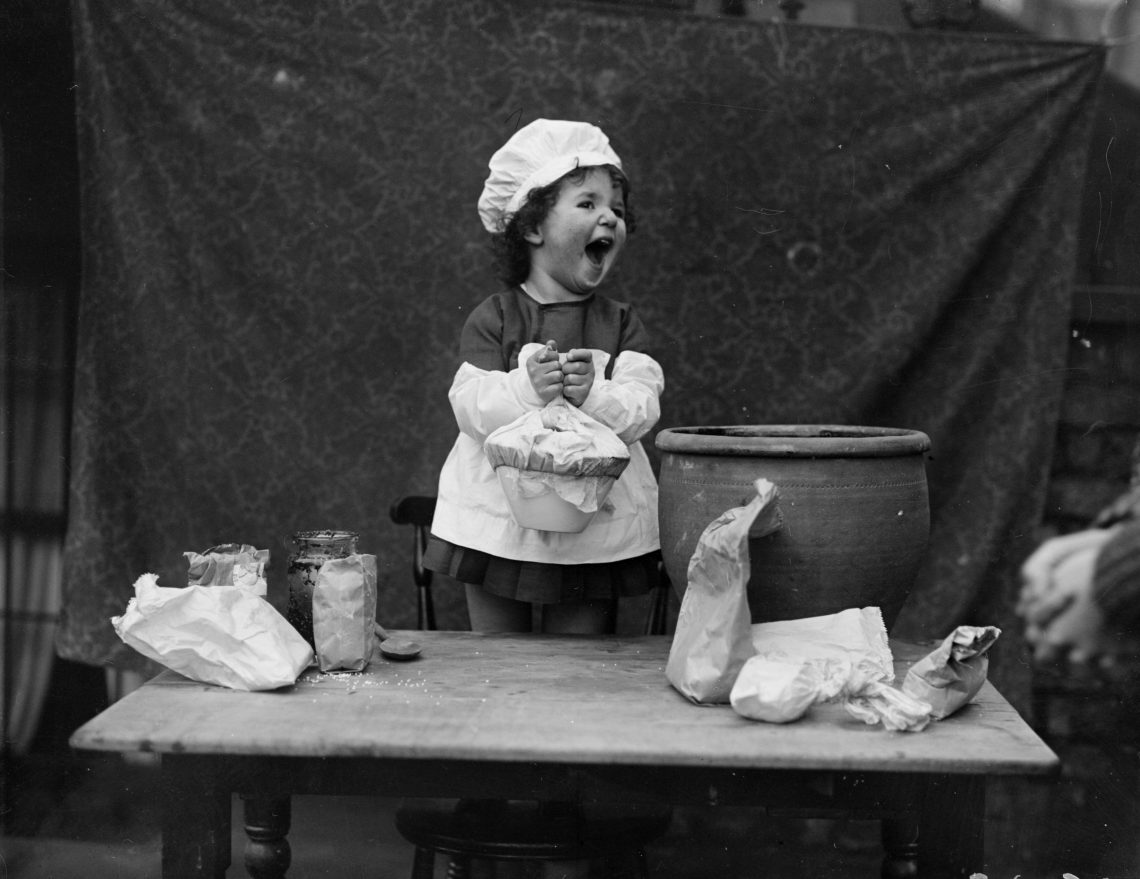 Ein kleines Mädchen mit Kochhaube steht lachend vor einer provisorischen Küche. Das Bild illustriert einen die Kolumne „Über das Lachen“ von Michael Köhlmeier.