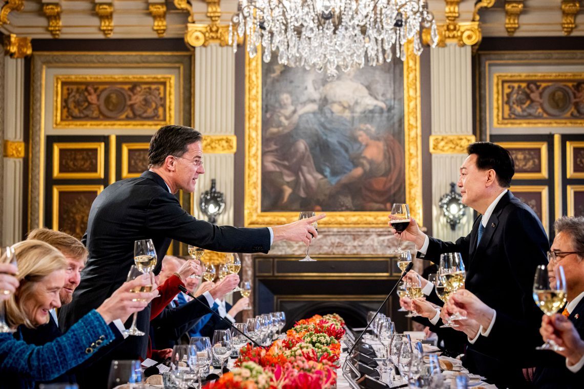 Der niederländische Ministerpräsident Mark Rutte prostet dem Präsidenten der Republik Korea Yoon Suk Yeol über einem prächtig mit Blumen geschmückten Tisch zu. Im Hintergrund sind Gemälde an tapezierten und goldbestuckten Wänden zu sehen. Das Bild illustriert einen Jahresrückblick.