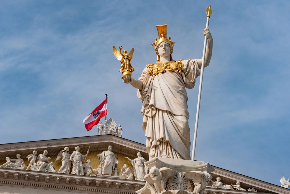 Foto einer Statue von Pallas Athene vor dem Parlamentsgebäude in Wien. Im Hintergrund vor strahlend blauem Himmel weht die Nationalflagge Österreichs.