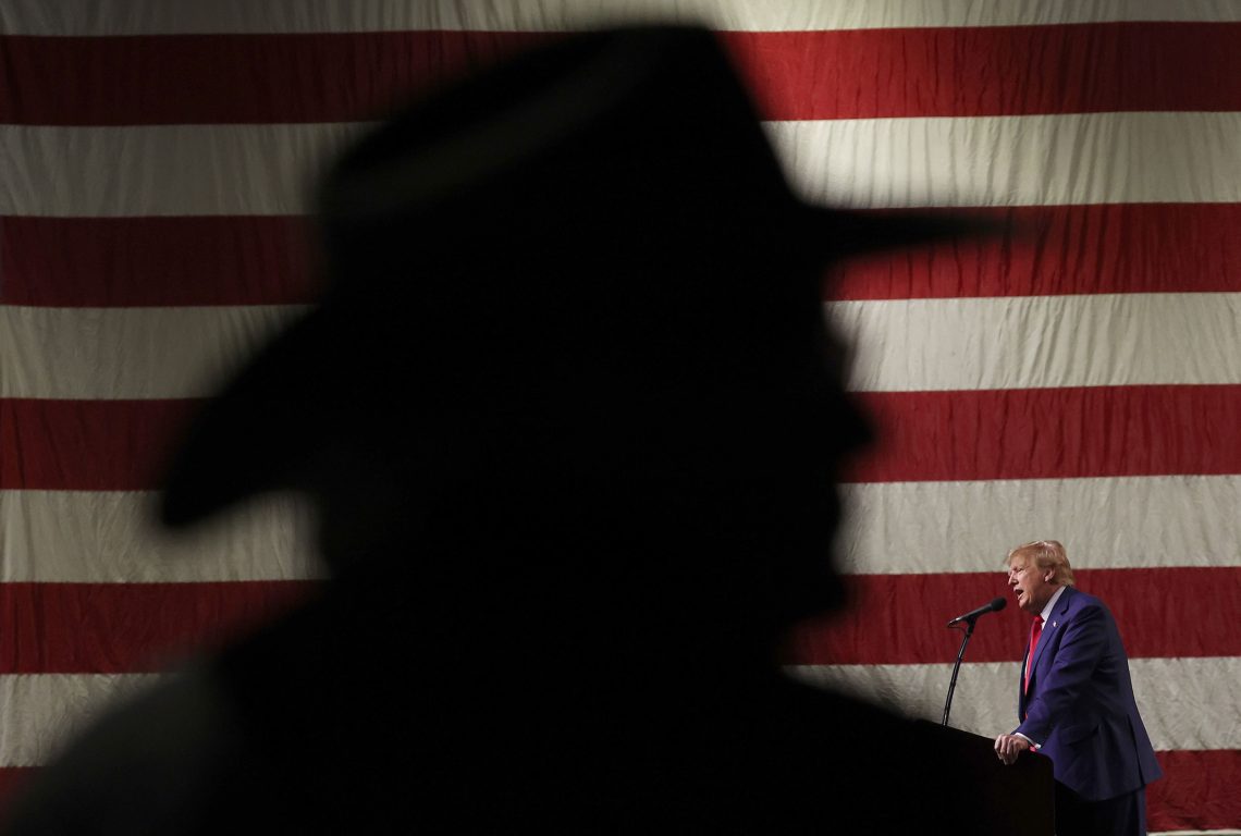 Donald Trump vor einer USA-Flagge neben dem gigantischen Schatten eines Cowboy-Hutes.
