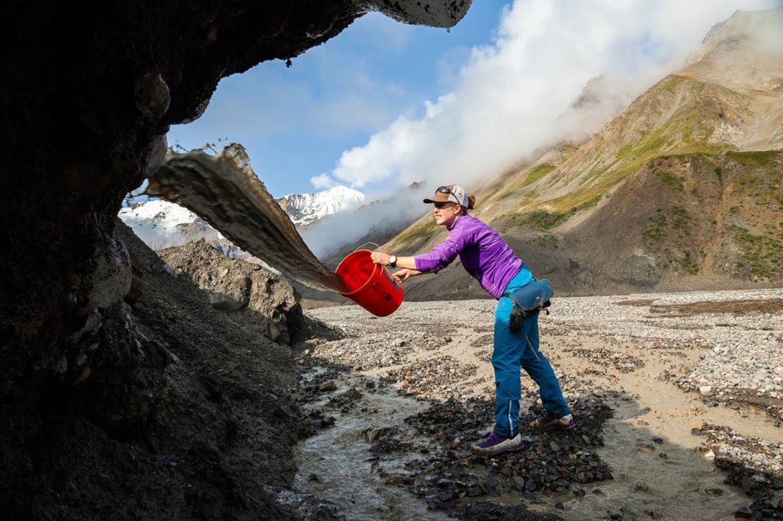 Mylène Jacquemart in Alaska bei der Vorbereitung von Forschung über Gletscher und Gletshertäler. Sie hält einen roten Eimer in der Hand und schüttet aus diesem eine Schlammmischung gegen eine Eiswand. Es ist der Rest eines Gletschers.