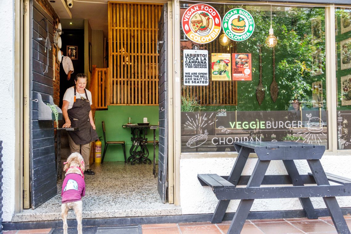 Ein Hund steht im Eingang eines veganen Restaurants und blickt auf die Besitzerin des Lokals, die ihn anlächelt.