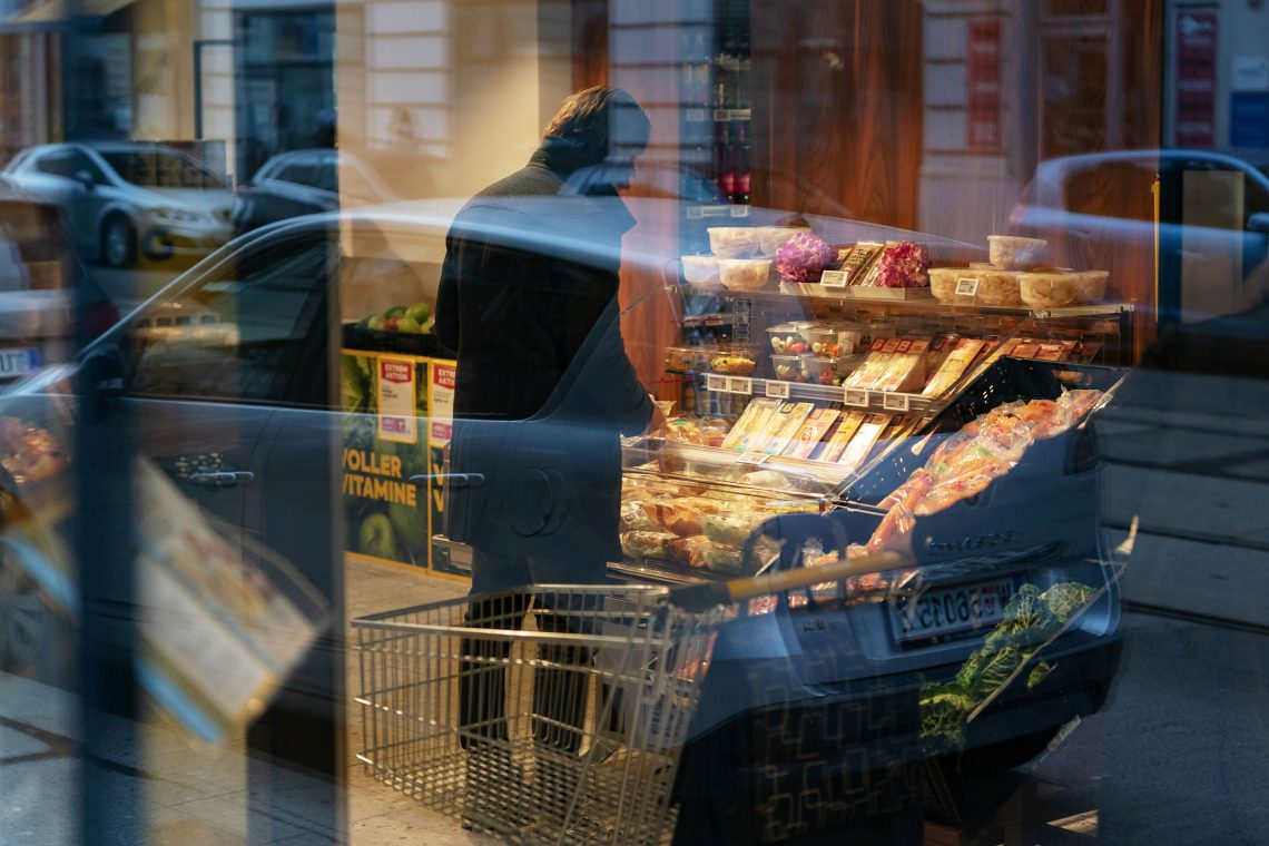 Durch sich spiegelnde Schaufensterscheiben sieht. man einen Mann, der in einem Supermarkt vor einer Auswahl Lebensmittel steht. Das Bild illustriert einen Beitrag über Inflationsraten im Vergleich.