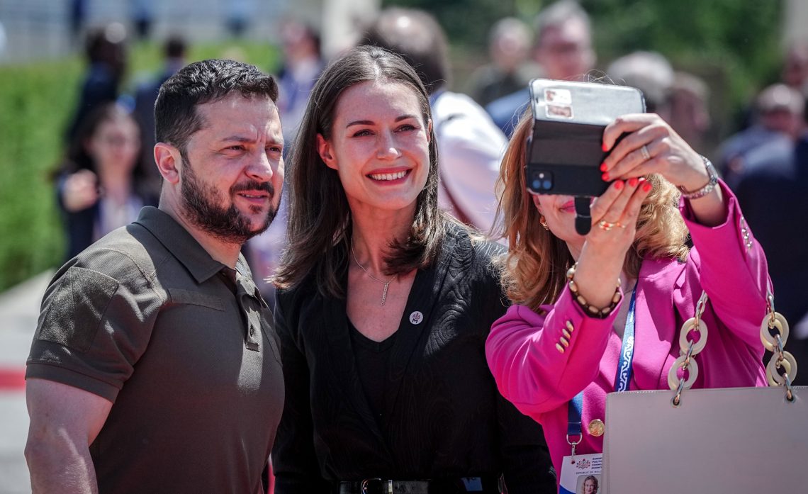 Wolodymyr Selensjij und Sanna Marin stehen neben einer Frau, die ein Selfie mit den beiden acht. Die Frau ist nicht zu erkennen, da ihr Gesicht vom Handy verdeckt wird.