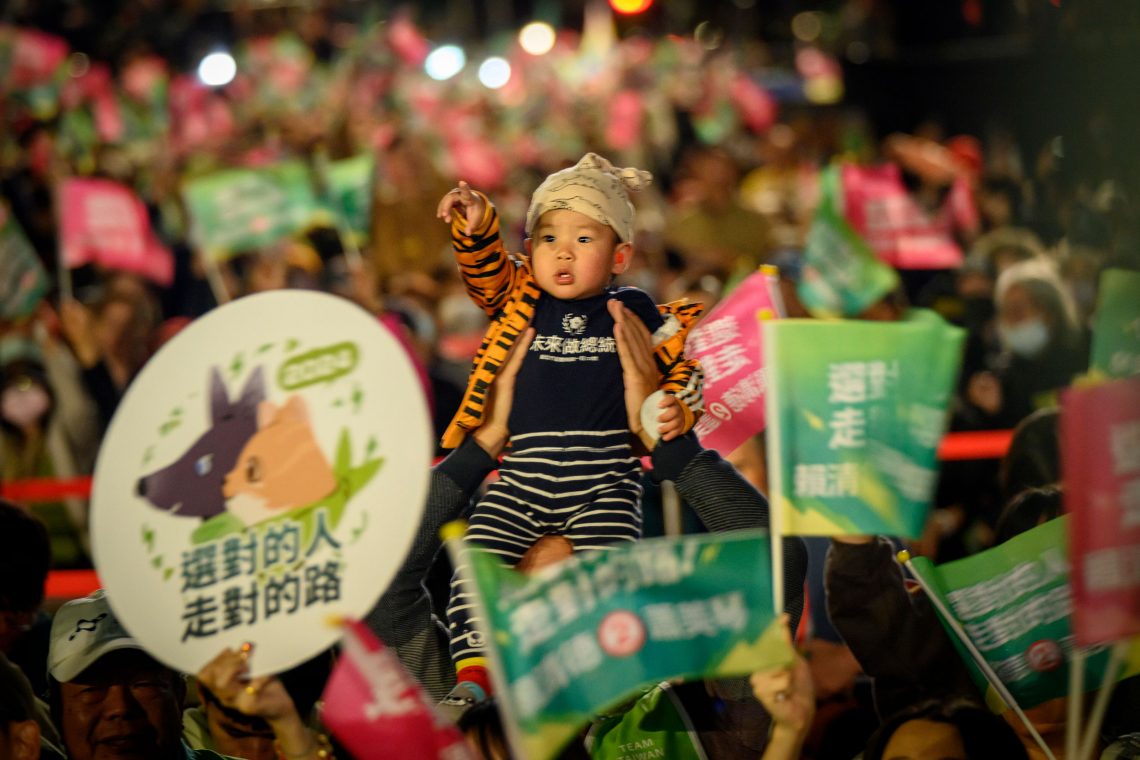 Ein kleines Kind in einem gesteiften Strampler wird in einer Menschenmenge auf den Schultern getragen und deutet Richtung Betrachter. Das Bild ist Teil eines Beitrags über Taiwan und die Wahlen.