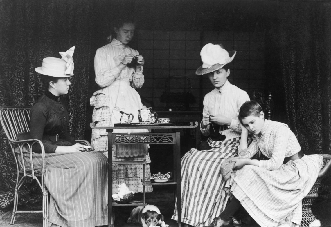 Schwarzweiß Fotografie von drei jungen Frauen einem Mädchen und einem Hund bei einer Tee_party Ende des 19. Jahrhunderts. Das Mädchen und der Hund unter dem Tee-Tischcen langweilen sich ostentativ. Das Bild ist Teil eines Beitrags zum Podcast Machthunger.
