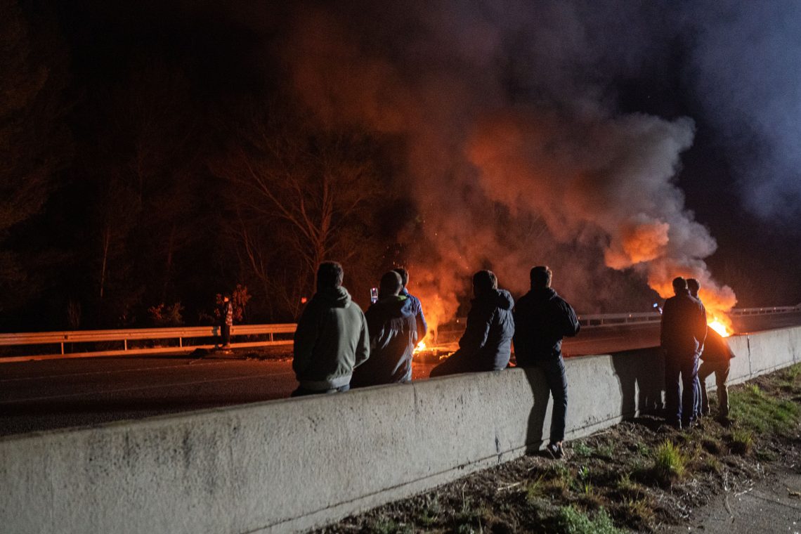 Feuer auf einer Autobahn. Mehrere Menschen stehen dabei und schauen auf die Flammen. Das Bild zeigt die Proteste von Bauern in Spanien im Februar 2024.