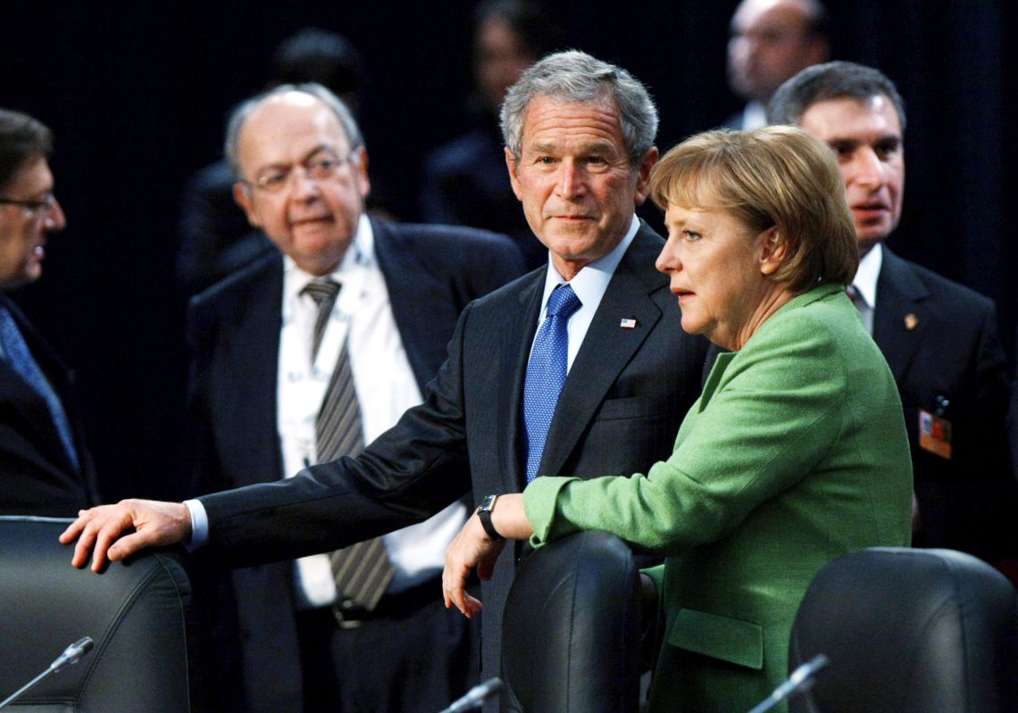 Angela Merkel und George W. Bush stehen nebeneinander und unterhalten sich wobei sie ihre Hnde auf die Rückenlehnen von Sesseln gestützt haben. Das Bild wurde vermutlich in einer Verhandlungspause aufgenommen. Es it Teil eines Beitrags über die Ursachen des aktuellen Ukraine-Kriegs.