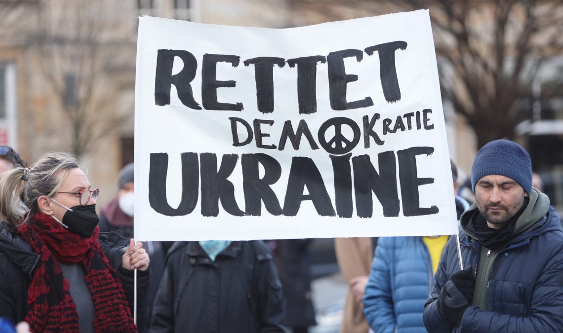 Teilnehmer einer Kundgebung für Frieden und gegen den russischen Angriffskrieg gegen die Ukraine stehen auf dem Alten Markt und halten ein Plakat mit der Aufschrift "Rettet die Demokratie Ukraine".