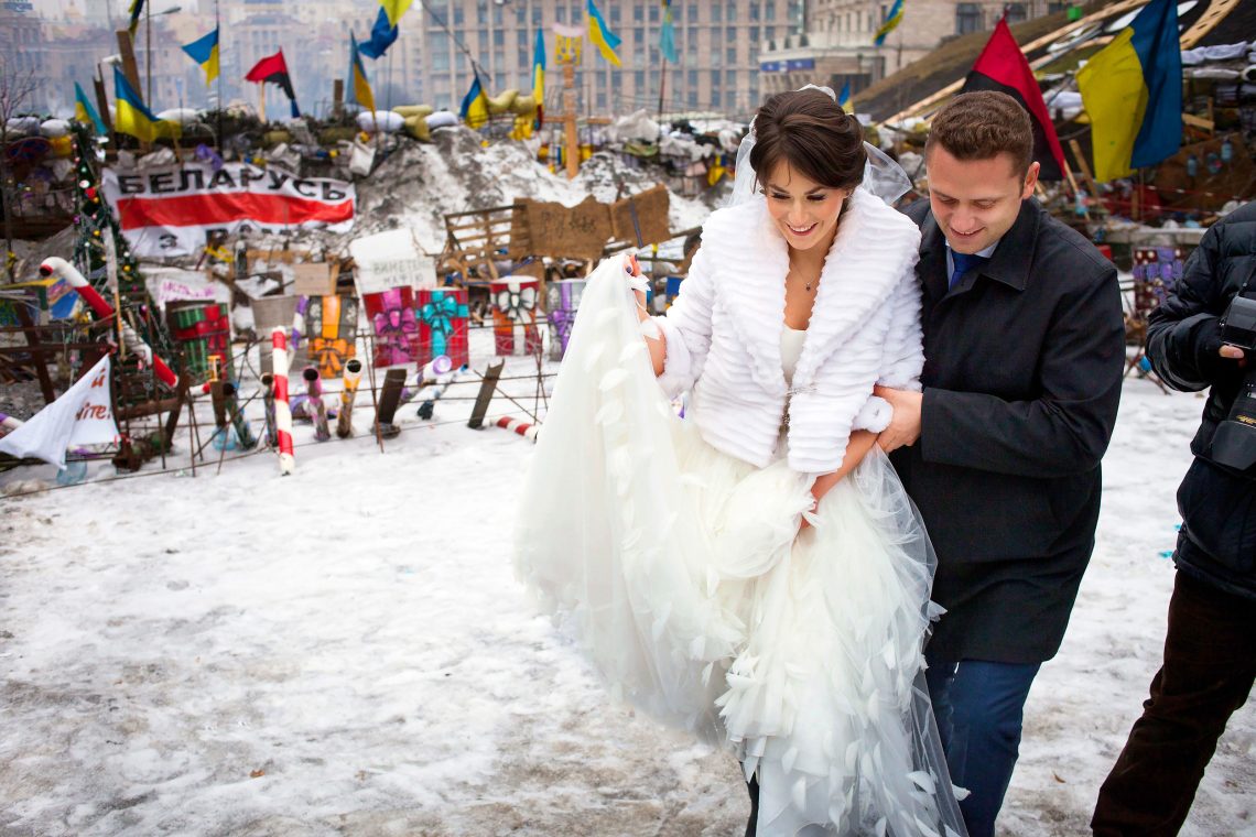Ein Hochzeitspaar auf dem Euromajdan in Kyjiw im Februar 2014. Es liegt Schnee und scheint kalt zu sein. Im Hintergrund Barrikaden und Fahnen.