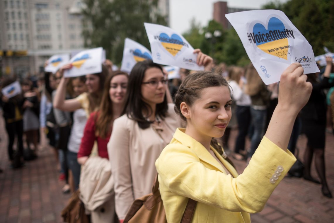 Eine Gruppe von jungen Frauen hält Zettel mit einem blau-gelben Herz auf dem Voice of Unity und 1944 und Vote for Ukraine steht, hoch. Daneben das Logo des Eurovision Song Contest.
