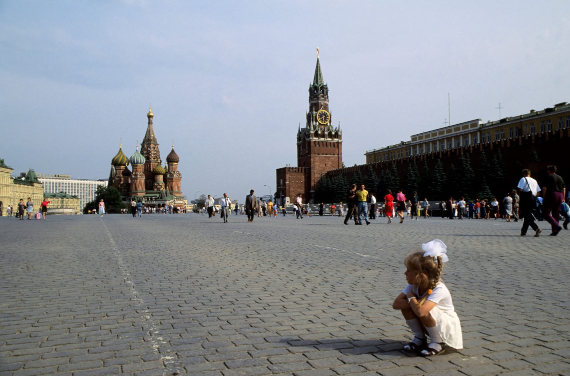 Ein Mädchen hockt auf dem roten Platz in Moskau in der Sonne. Im Hintergrund ist der Kreml zu sehen.
