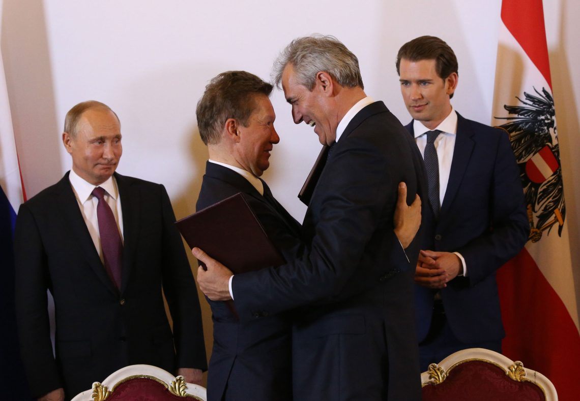 Alexei Miller und Rainer Seele umarmen sich und blicken einander in die Augen. Im Hintergrund Wladimir Putin und der damalige österreichische Bundeskanzler Sebastian Kurz neben einer Österreich-Fahne. Rainer Seele hält einen Vertrag in den Händen.