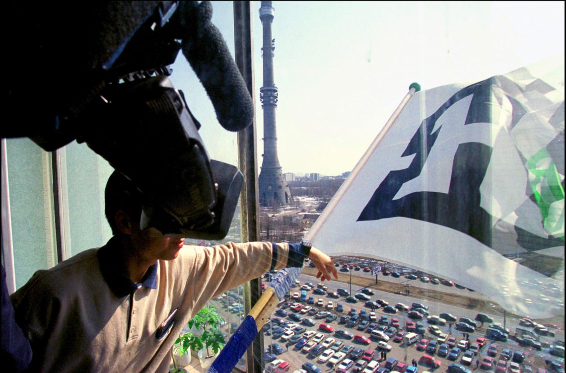 Ein Mann schwenkt eine große Fahne aus einem Gebäude. Neben ihm steht eine Kamera. Unten ist ein Fernsehturm und ein großer Parkplatz zu sehen.