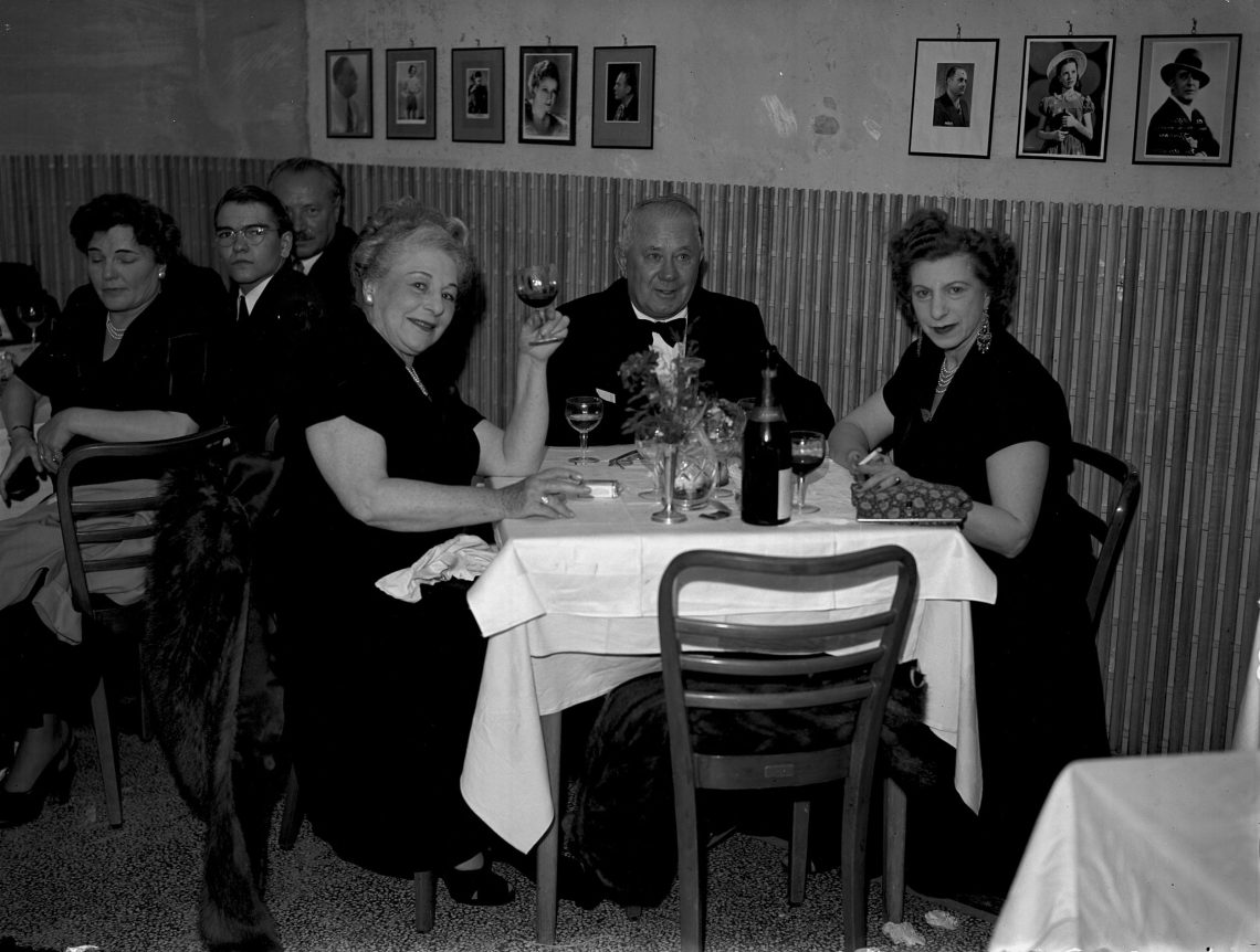 Drei Personen, zwei Frauen und ein Mann, sitzen festlich gekleidet an einem Tisch in einem Restaurant. Eine der beiden Frauen prostet dem Betrachter zu, die andere blickt ernst in die Kamera. Beide Frauen rauchen. Aug dem Tisch steht eine Flasche Wein. Das Bild illustriert einen Beitrag über Salz.