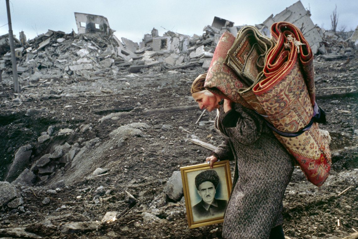 Eine Frau trägt Teppiche auf dem Rücken und hält in der rechten Hand ein goldgerahmtes Portrait eines Mannes. Sie geht durch Ruinen.