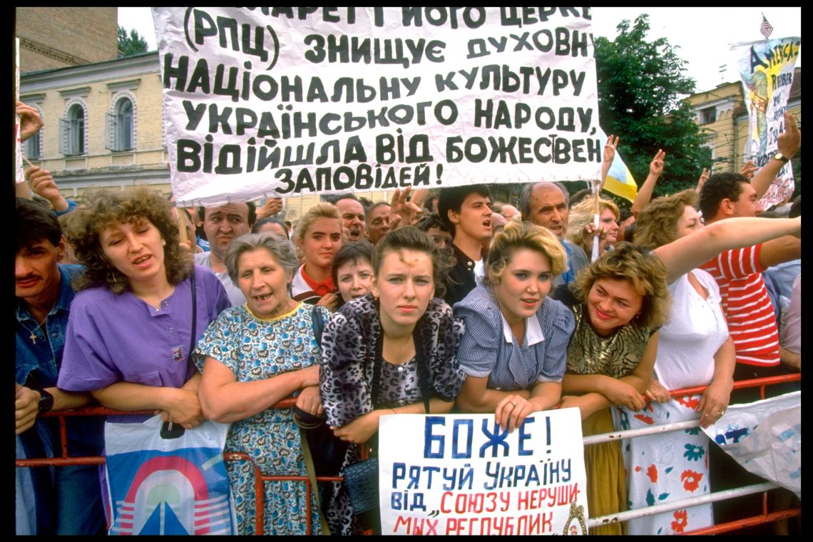 Demonstranten und Demonstantinnen mit Plakaten stehen an einer Straßenabsperrung und winken und skandieren etwas. Das Bild illustriert einen Beitrag über die Ursachen des Ukraine-Krieges.