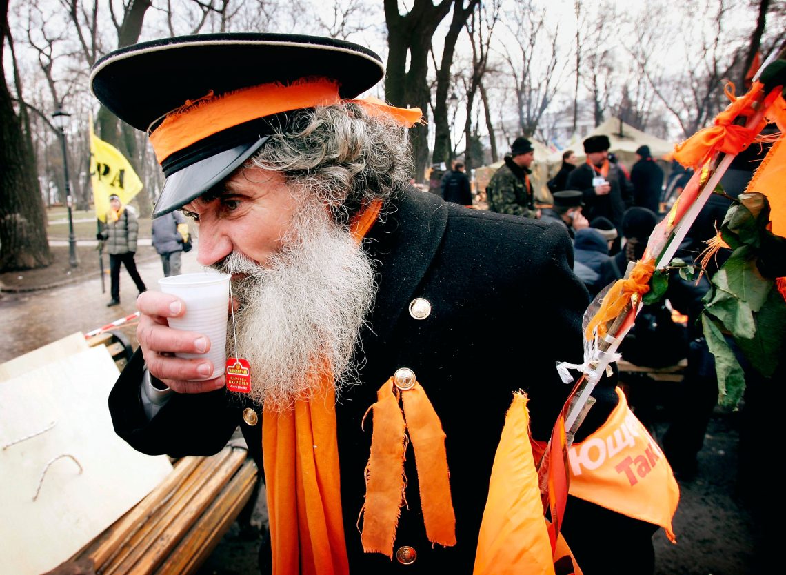 Ein Mann in einer Fantasieuniform mit orangen Schleifen und Bändern steht im Winter auf einem Platz mit anderen Demonstrierenden und trinkt aus einem Plastikbecher warmen Tee.