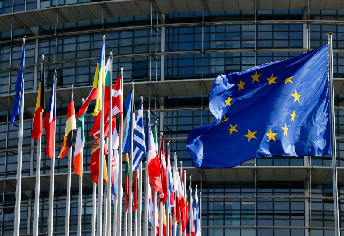 Das Bild zeigt im Vordergrund die Europa-Flagge und links daneben die Flaggen der Mitgliedsländer vor dem Louise-Weiss-Gebäude des Europäischen Parlaments in Straßburg. Das Bild illustriert einen Kommentar über Europas Schlagkraft.