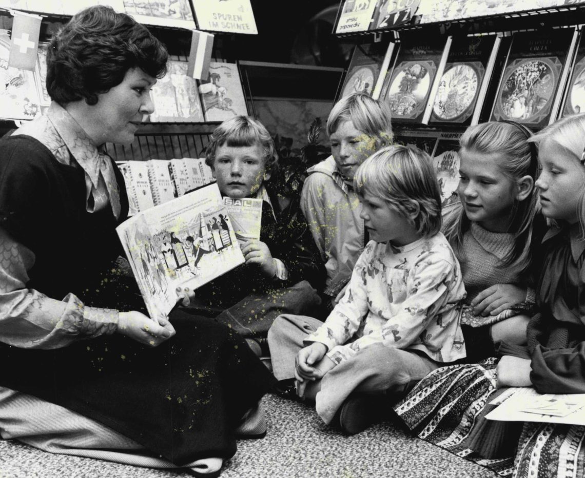 Eine Frau erzählt in einer Bücherecke fünf Kindern eine Geschichte aus einem aufgeschlagenen Buch. Das Bild illustriert einen Artikel zum Thema, was gute Erzähler ausmacht.