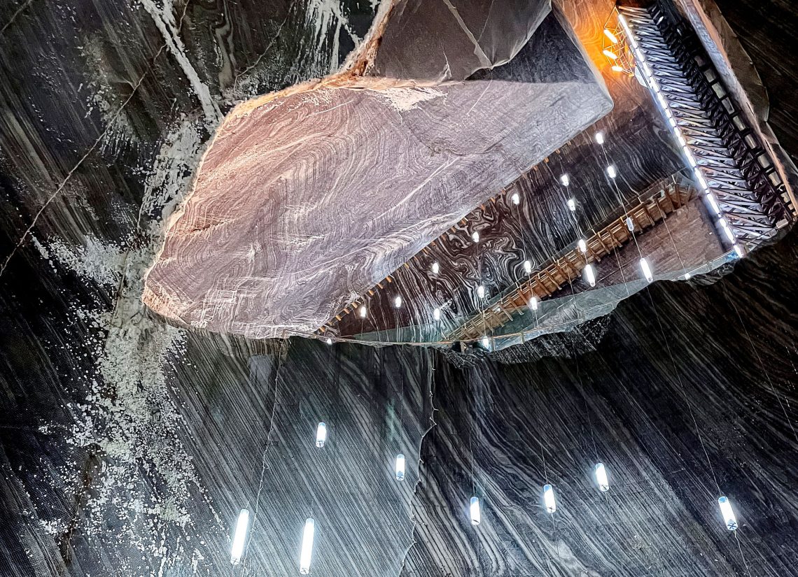 Der Innenraum der Salzkaverne im Bergwerk Turda in Rumänien wird von Hängeleuchten erstrahlt. Die Wände zeigen eine grün-weiße Maserung. Eine Zugangsloch bietet eine Besuchergalerie für Touristen. Das Bild illustriert einen Beitrag über die Rolle von Salz für die Wirtschaft.