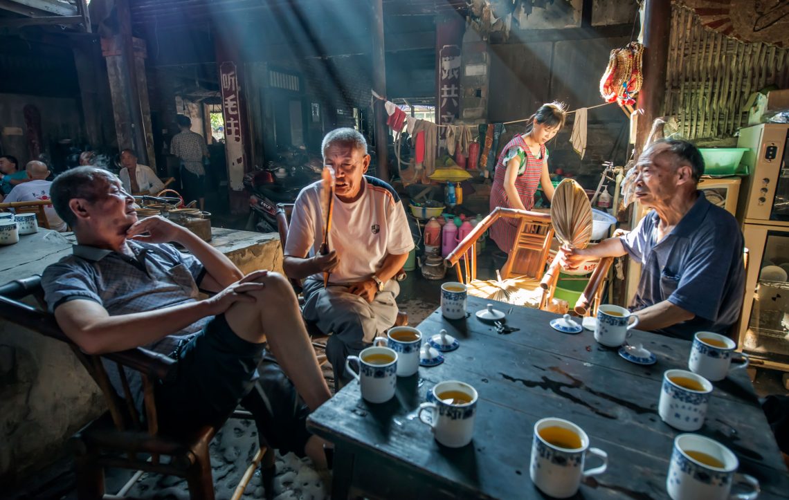 Auf einem großen Tisch, an dem drei Männer sitzen und reden, stehen viele Tassen mit Tee. Durch ein Oberlicht fällt Sonne in den Raum. Eine Frau steht im Hintergrund und arbeitet.