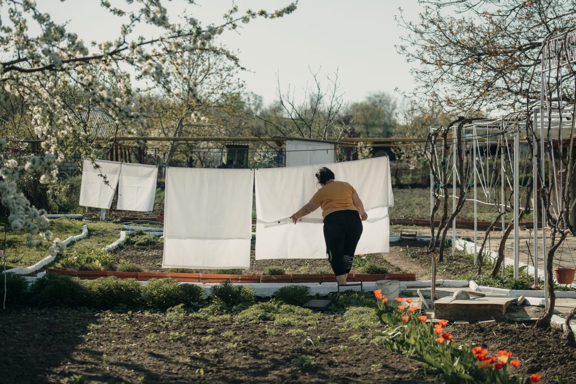 In Gagausien, Moldau, streicht eine Frau ein weißes Betttuch glatt, das neben einem Laken und zwei Kopfkissen an einer Leine in einem Garten mit blühenden Obstbäumen hängt.