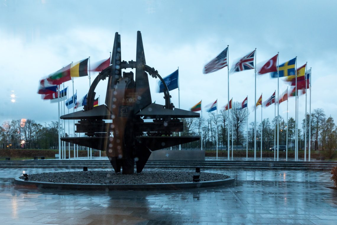 Der NATO-Stern ist durch ein Fenster mit Regentropfen im NATO-Hauptquartier vor den Flaggen der 32 Mitgliedsstaaten zu sehen. Das Bild illustriert einen Kommentar über Österreichs Neutralität und warum die EU das Kielwasser der NATO verlassen sollte.