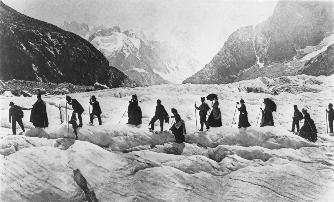Eine Gruppe von Männern und Frauen spaziert mit Sonnenschirmen und Stöcken ausgestattet auf einem Gletscher. Die Frauen tragen lange Kleider und die Männer Hut und Gehrock. Das Foto stammt aus dem 19. Jahrhundert und zeigt eine Form des frühen Alpentourismus.