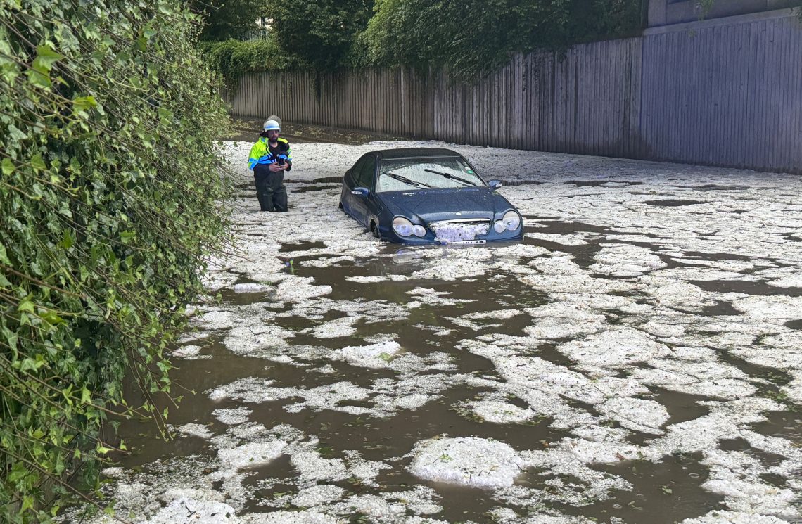 Eine Unterführung mit einem Auto, dass in Wassermassen eingeschlossen ist. Auf der Wasseroberfläche schwimmt Hagel. Das Bild illustriert einen Beitrag über Klimawandel.