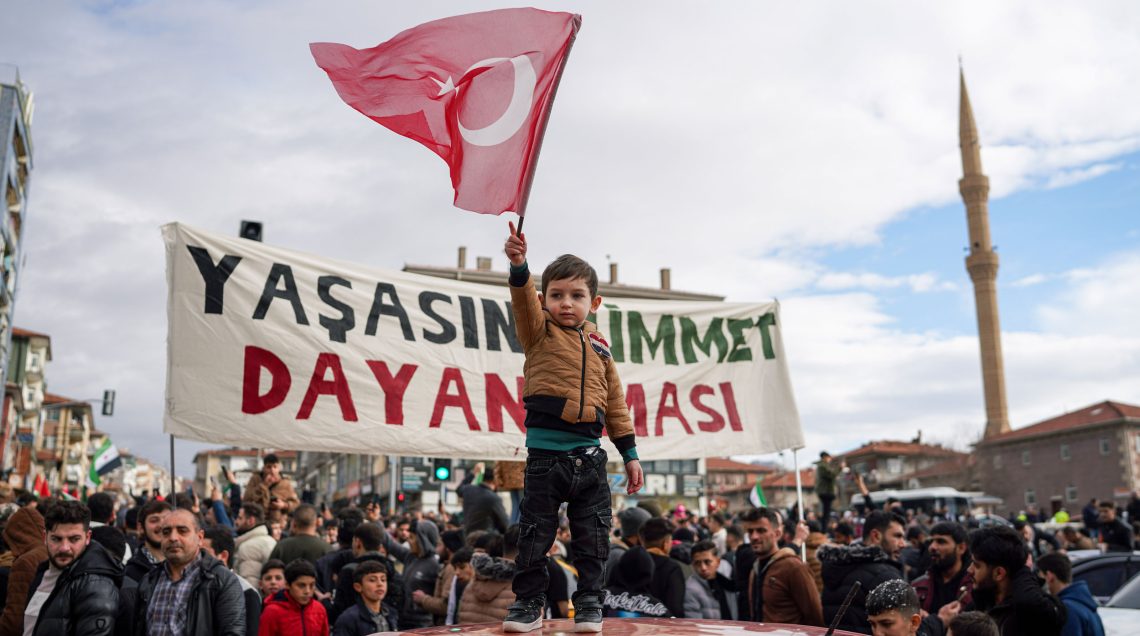 Bild zum Thema Türkei in Syrien: Ein syrisches Kleinkind steht auf einem Auto auf den Straßen Ankaras und schwenkt eine türkische Fahne umringt von feiernden Syrern.
