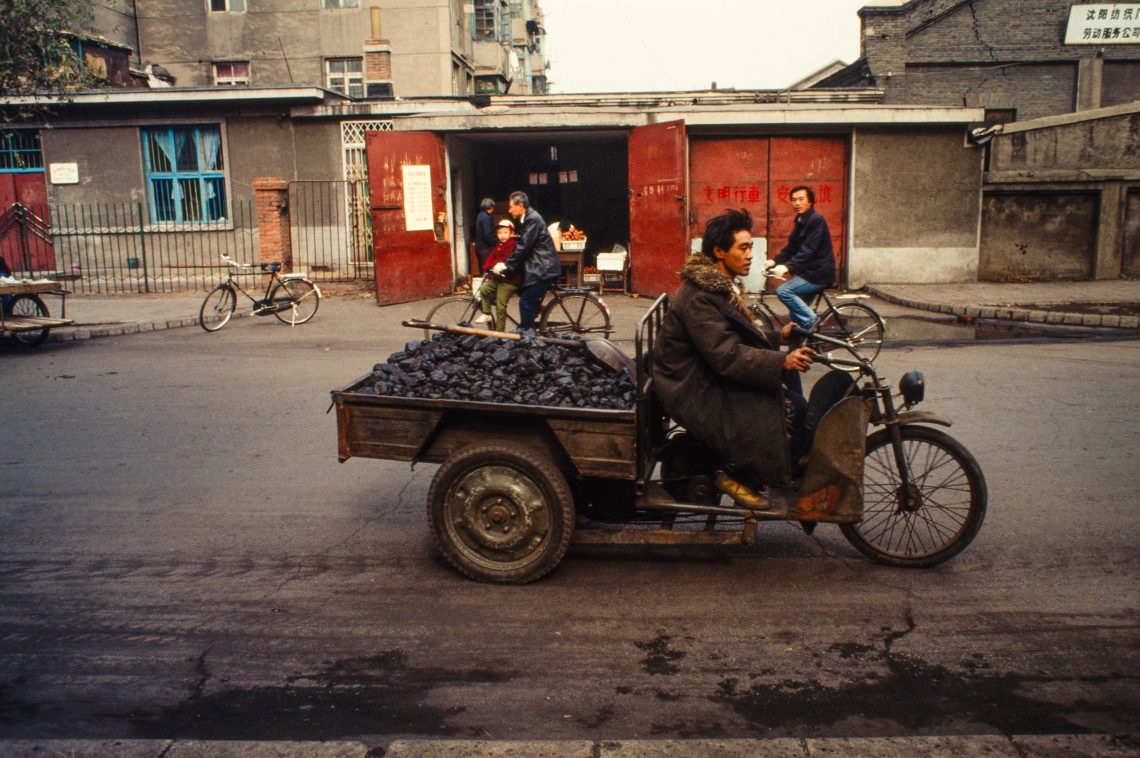 Ein Kohletransport mit einem Lasten-Motorrad in Shenyang im Nordosten Chinas. Im Hintergrund ist ein Verkaufsladen zu sehen und eine Häuserzeile mit grauem Verputz. Einige Fahrradfahrer sind zu sehen. Das Bild ist Das Bild ist Teil eines Podcasts mit Susanne Weigelin-Schwiedrzik, in dem sie unter anderem über die Wiederbelebung des Staatskapitalismus durch Xi Jinping spricht. Die Wirtschaft Chinas wächst nicht mehr im selben Ausmaß wie noch vor einigen Jahren. Dies hat, so Weigelin-Schwiedrzik, auch mit der Zurückdrängung der Privatwirtschaft und dem Niedergang des informellen Sektors zu tun. Während die Exportzahlen weiterhin gut seien, schrumpften die Erlöse aus diesem Sektor.