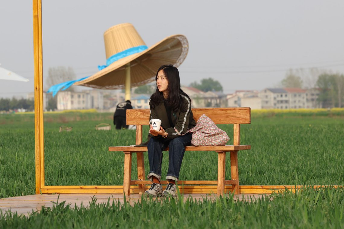 Eine Frau sitzt mit einem Becher Kaffee unter einem großen Hut auf einer Bank. In einem Café „auf dem Land“ in der Nähe von Xiangyang (Hubei). Cafés wie dieses inmitten von Feldern sind beliebt. Mit dem gestiegenen Wohlstand einer meist urbanen Mittelschicht steigt die Sehnsucht nach dem „einfachen“ Landleben. Viele Bauern leben in China – trotz weit fortgeschrittenen Technologien wie etwa drohnenunterstützte Ernten in großer Armut. Das Bild ist Teil eines Podcasts mit Susanne Weigelin-Schwiedrzik, in dem sie unter anderem über die Wiederbelebung des Staatskapitalismus durch Xi Jinping spricht. Die Wirtschaft Chinas wächst nicht mehr im selben Ausmaß wie noch vor einigen Jahren. Dies hat, so Weigelin-Schwiedrzik, auch mit der Zurückdrängung der Privatwirtschaft und dem Niedergang des informellen Sektors zu tun. Während die Exportzahlen weiterhin gut seien, schrumpften die Erlöse aus diesem Sektor.