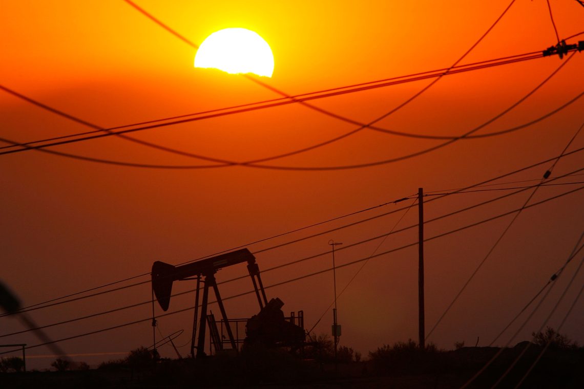 Das Bild zeigt eine Szene bei Sonnenuntergang. Im Vordergrund ist eine Silhouette einer Ölpumpe zu sehen, die von mehreren Stromleitungen und Masten eingerahmt wird. Das Bild illustriert einen Artikel über Klimaökonomie.