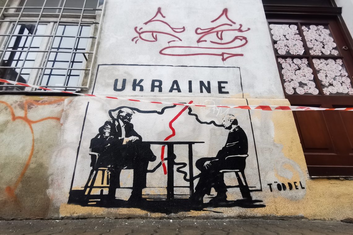 Das Bild zeigt ein Graffiti mit der Aufschrift Ukraine und Trump und Putin, die an einem Tisch sitzen. Im Hintergrund ist eine Karte der Ukraine zu sehen, die durch eine rote Linie getrennt ist. Das Bild illustriert einen Artikel über die Lehren aus dem Krieg in der Ukraine.