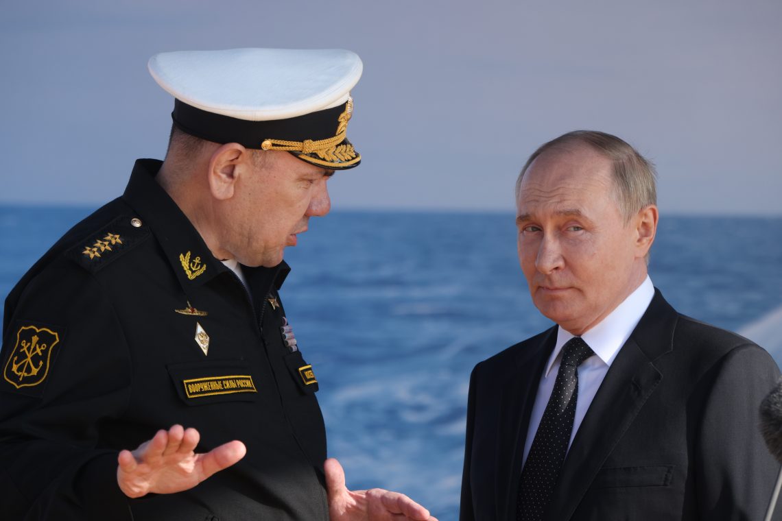 Der russische Präsident steht mit dem Oberbefehlshaber der russischen Marine an einer Meereskulisse und spricht mit ihm, während beide Uniform beziehungsweise Anzug tragen. Im Hintergrund ist das Wasser zu sehen; die Szene findet bei einem Besuch des Sevmash‑Schiffbauwerks in Sewerodwinsk am 24. Juli 2025 statt.