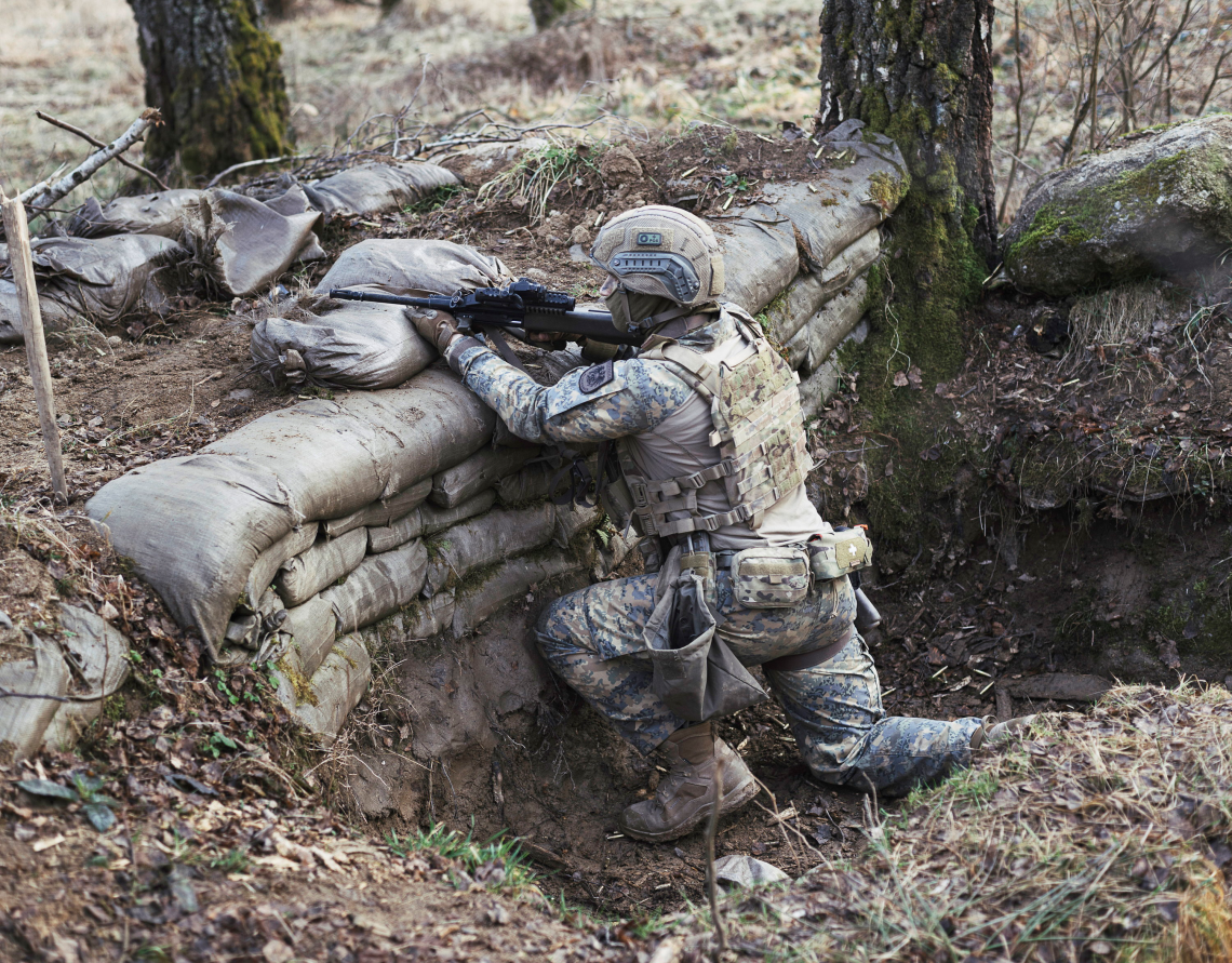 Bild zum Thema Neutralität: Eine aus Erde und Sandsäcken gebaute Stellung im Freien. Eine Person in Tarnkleidung und Schutzausrüstung kniet hinter der Sandsackwand und hält ein Gewehr in Richtung des offenen Geländes. Der Boden ist uneben und bewachsen, und im Hintergrund stehen mehrere Bäume.